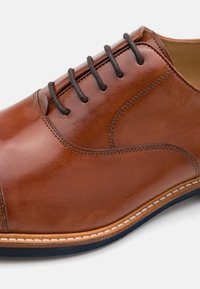 Zapato de cuero marrón con una textura lisa, punta redonda y cordones oscuros. Presenta un pespunte claro en contraste a lo largo de la suela.