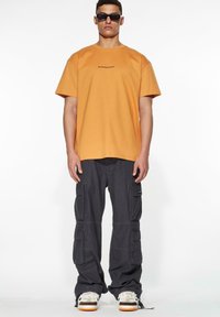 Orange bomulds T-shirt med korte ærmer og sort logo; parret med mørkegrå cargo-bukser med flere lommer og brede ben.