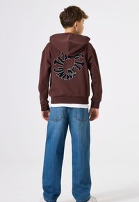 Sudadera con capucha marrón con un diseño gráfico de "TEAM" en negro en la parte posterior. Combinada con vaqueros azules holgados y zapatillas blancas.