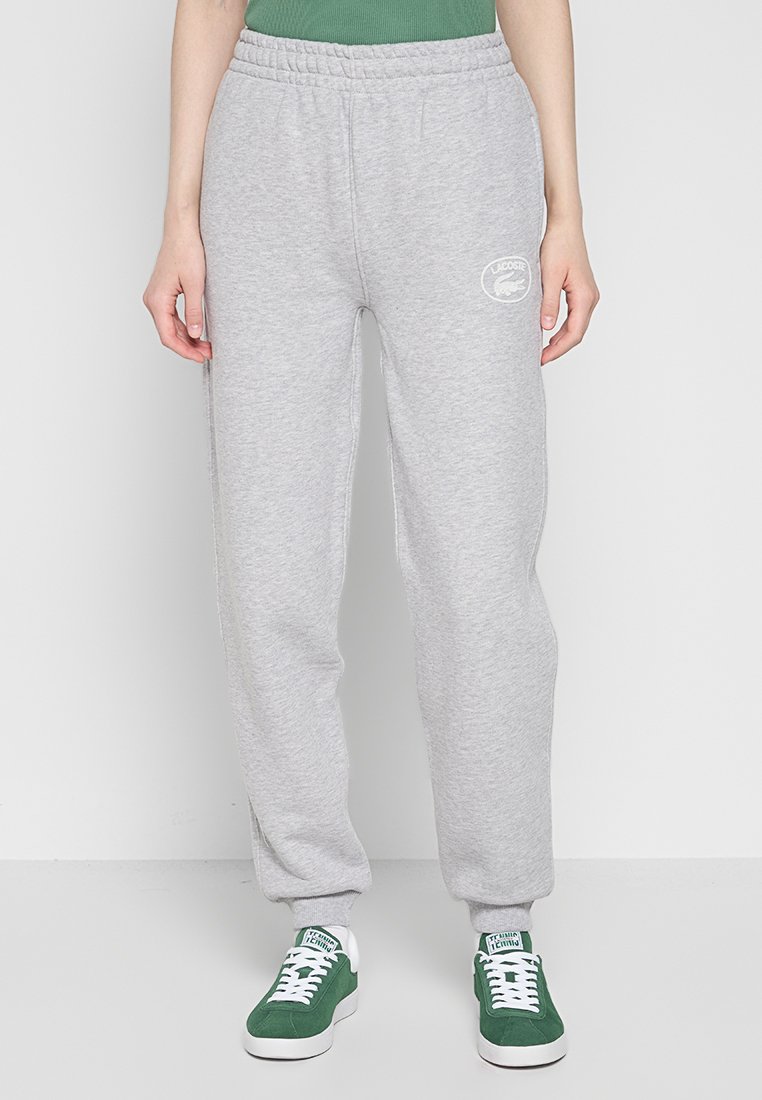 Osoba w jasnoszarych dresach Lacoste i zielonych trampkach z białymi sznurówkami, stojąca na jednolitym białym tle.