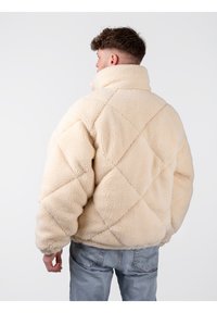 Chaqueta de forro polar sherpa en color beige con patrón de rombos acolchado, cuello alto y puños elásticos, que presenta un corte relajado y una textura suave.