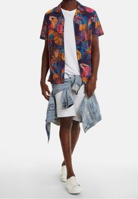 Camicia a maniche corte con sfondo blu scuro e motivo di foglie tropicali colorate. Indossata sopra una t-shirt bianca e abbinata a pantaloncini di denim.