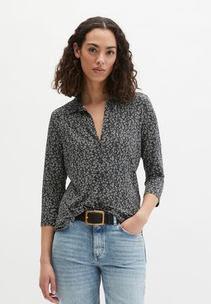 Frau mit lockigem Haar, die ein schwarzes, geblümtes Hemd mit Knöpfen, hellblaue Jeans und einen schwarzen Gürtel mit einer goldenen, rechteckigen Schnalle trägt.