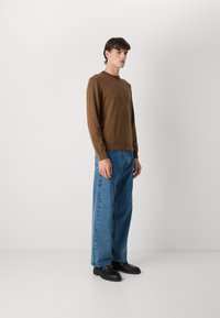 Bruine getailleerde trui, blauwe wijde jeans en zwarte loafers. De stoffen zijn glad, met een strak ontwerp en minimale stikdetails.