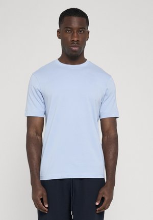 Modelo masculino vistiendo una camiseta azul claro de manga corta y pantalones oscuros, de pie sobre un fondo blanco liso.