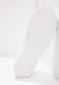 White sneaker sole with star-patterned tread resting against a white corner surface, showing the bottom texture and edge details.