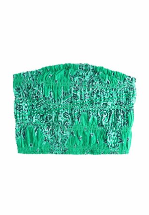 Frau mit langen braunen Haaren, die ein grünes gemustertes Tubetop und passende Hosen trägt, steht seitlich vor leuchtend pinken Blumen.