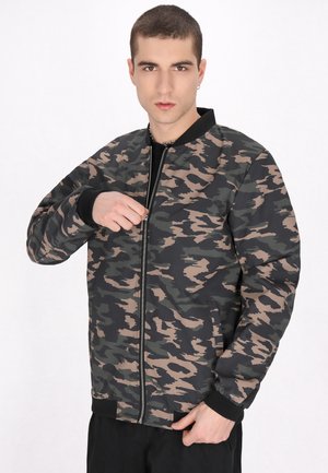 Jonge man met kort haar die een zwart-bruin camouflage ritsjack draagt tegen een effen witte achtergrond.