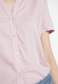 Blouse rose clair à manches courtes avec un col en V, petits pois blancs, et six boutons assortis sur le devant. Tissu doux et légèrement texturé.