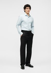 Bărbat tânăr stând cu mâinile în buzunare, purtând o cămașă albastru deschis cu nasturi, pantaloni negri și pantofi negri eleganți, pe un fundal alb simplu.