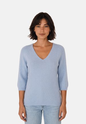 Mujer con cabello corto y oscuro que lleva un suéter de cuello en V azul claro con mangas tres cuartos y vaqueros azul claro, de pie frente a un fondo liso.