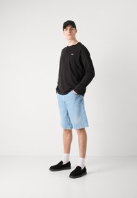 Juodas ilgomis rankovėmis marškinėliai, šviesiai mėlyni džinsiniai šortai ir juodi slip-on batai su baltais kojinėmis. Modelis stovi pasitikėdamas savimi, rankas kišenėse.