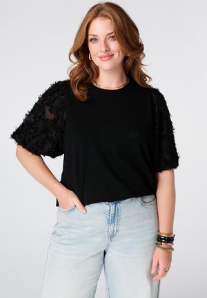 Vrouw met golvend rood haar die een zwart topje met gestructureerde pofmouwen draagt en lichtblauwe spijkerbroek, glimlachend met één hand in de zak.