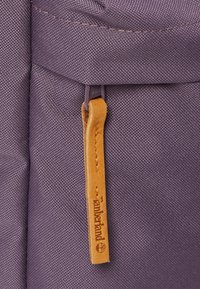 Lila tygryggsäck med en texturerad yta, en zippered ficka med en dragflik i ljust läder och en framträdande logotypdetalj.