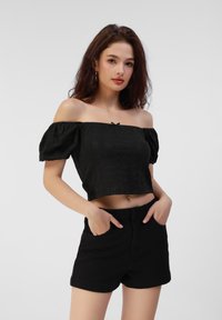 Sort off-the-shoulder cropped top med puffede ærmer, featuring øjelet tekstur, og en front knude detalje, parret med højtaljede sorte shorts.
