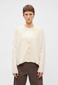 Ανοιχτό κρεμ πουλόβερ φτιαγμένο από μαλακό ύφασμα, με V-λαιμόκοψη, πέντε κουμπιά, ρίχτες ώμους και ελαφρώς κοντή, άνετη εφαρμογή.