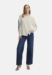 Camisa blanca de botón con bordado floral azul en las mangas, combinada con jeans de tiro alto de pierna ancha en azul oscuro y tacones negros con tiras.