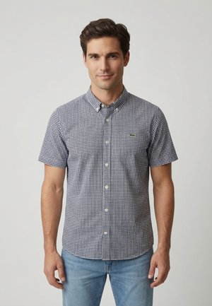 Hombre de pie contra un fondo liso, vistiendo una camisa de cuadros en blanco y negro de manga corta con botones y jeans azul claro.