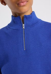 Blauwe gebreide pullover met een hoge kraag en een zilveren rits. De stof heeft een gladde textuur en het ontwerp is figuurcorrigerend.