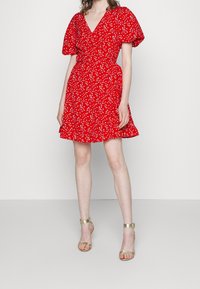 Vestido rojo ajustado con mangas cortas abullonadas, con patrones florales blancos. Falda acampanada y diseño de escote en V. Llevado con sandalias doradas de tiras.