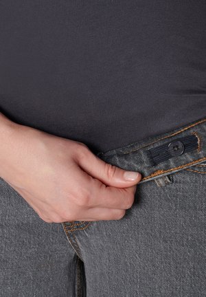 Grijze denim jeans met oranje stiksels, voorzien van een knoopsluiting. Een hand grijpt de tailleband, waardoor de stoftextuur zichtbaar wordt.