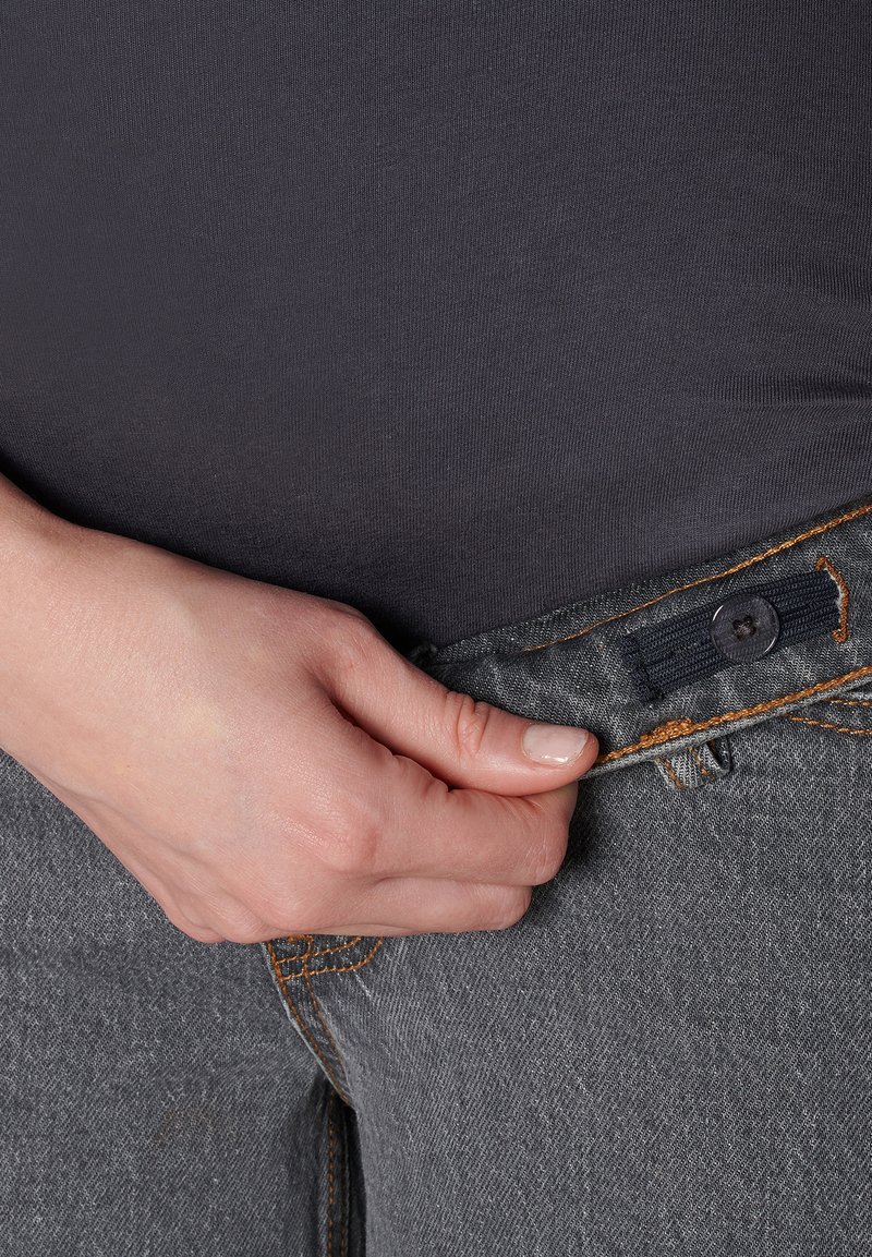 Grå denimjeans med orange sömmar, med en knappstängning. En hand greppar midjebandet och visar tygtexturen.
