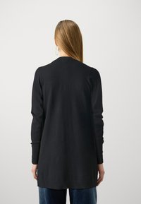Svart långärmad cardigan med avslappnad passform, rak skärning, ribbade mudd och fåll, tillverkad av ett mjukt material, bärs över blå jeans.