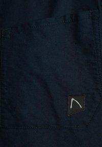 Bolsa de tela azul marino con un pequeño parche negro que presenta un gráfico blanco. Superficie texturizada con costuras limpias a lo largo de los bordes.