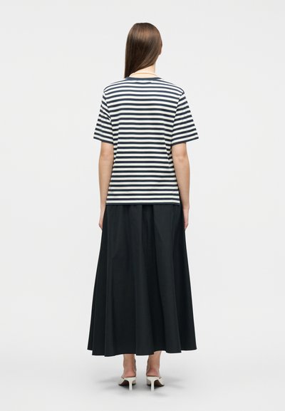 Γυναίκα με την πλάτη, φορώντας μπλούζα με κοντά μανίκια σε ναυτικό μπλε και λευκές ρίγες, μακριά μαύρη φούστα, λευκά σανδάλια με τακούνι και κολιέ με μαργαριτάρια.