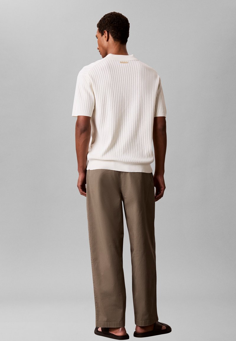 Homme de dos portant un pull blanc court à manches courtes en maille côtelée, un pantalon large marron et des sandales noires sur un fond gris.