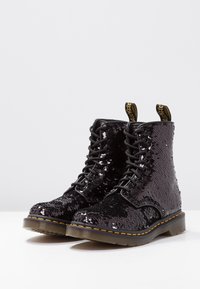 Botas de tobillo negras con lentejuelas, diseño de cordones, parte superior texturizada y tiradores amarillos con logo. Presenta una suela robusta y costuras en contraste.