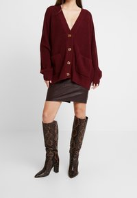 Femme portant un cardigan bordeaux à boutons, une mini-jupe en cuir marron foncé et des bottes en cuir serpent haute jusqu'aux genoux.
