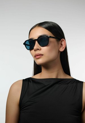 Lunettes de soleil noires avec une forme géométrique et des verres bleu foncé. Présente une monture noire mate avec un branding subtil sur les branches.
