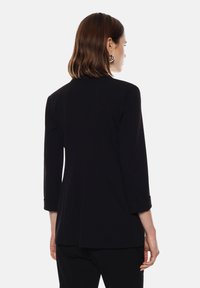 Donna con capelli castani di lunghezza spalle, indossa orecchini a cerchio d'argento e un blazer nero a maniche lunghe, vista da dietro.