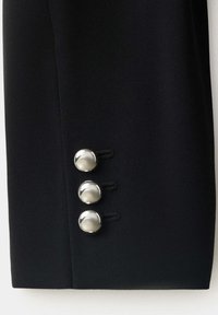 Gros plan sur la manche noire d'une veste avec trois boutons ronds argentés brillants disposés verticalement près du poignet.