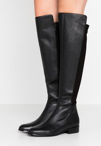 Botas de piel negra hasta la rodilla con un acabado suave y cremallera lateral; presentan paneles de ante para añadir textura y un tacón bajo y cuadrado.