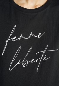 Camiseta negra de cuello redondo con texto en cursiva blanca que dice "femme liberté". El material parece suave y la tela tiene una textura sutil.