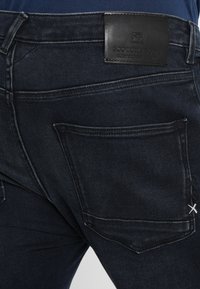 Dunkelblaue Jeans mit einer Nahaufnahme der Gesäßtasche. Merkmale sind ein lederner Bund-Patch und subtile Stecknähte. Glatte Textur.