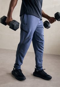 Pantalons de sport gris avec une taille ajustable, poche zippée sur le côté et accents noirs, associés à des chaussures de sport noires, tenant des haltères.