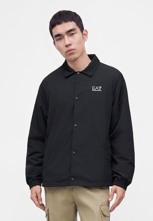 Young man wearing a black EA7 buttoned jacket and beige cargo pants standing against a plain light background.