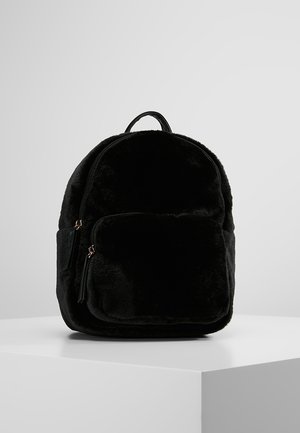 Zwarte pluche rugzak met voorvak met ritssluiting en bovenste handvat, tentoongesteld op een wit voetstuk tegen een grijze achtergrond.