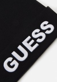Bonnet noir avec le texte "GUESS" brodé en blanc. Tissu tricoté doux, forme arrondie et texture lisse. Pas de motifs ou d'accents supplémentaires.