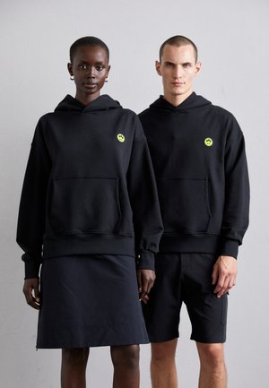 Schwarze Hoodies mit Fronttaschen, featuring ein kleines neon grünes Logo, über dunkler Kleidung getragen. Glatter Stoff, entspannter Schnitt, minimalistisches Design.