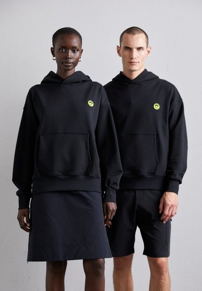 Hanorace negre cu buzunare frontale, având un mic logo neon verde, purtate peste îmbrăcăminte închisă la culoare. Material neted, croială lejeră, design minimalist.