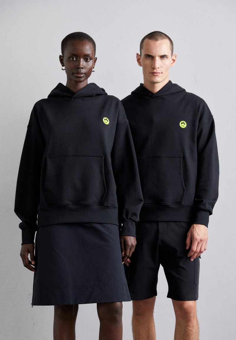 Black hoodies with front pockets, featuring a small neon green logo, worn over dark clothing. Smooth fabric, relaxed fit, minimal design.