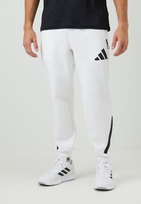 Bílé sportovní kalhoty s černými akcenty, se širokým elastickým pasem, boční kapsou na zip a strukturovaným designem. V kombinaci s bílými teniskami.