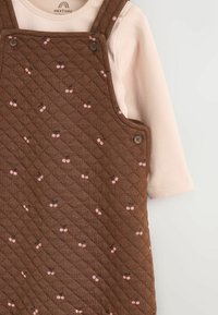 Brązowe pikowane ogrodniczki z drobnym różowym wzorem wisienek, ozdobione brązowymi guzikami. Pod spodem widać jasno-różową bluzkę z długim rękawem.