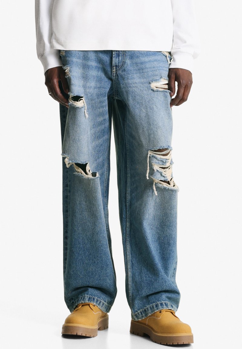 Person wearing light blue ripped jeans with large tears and tan work boots, standing against a white background.