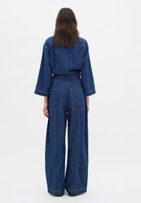 Jeansoverall in Dunkelblau mit weitem Bein, langen Ärmeln und sichtbaren Nähten. Verfügt über zwei große Gesäßtaschen und eine taillierte Taille.