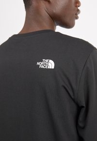 Schwarzes Langarmshirt aus weicher Baumwolle mit einem weißen Logo im oberen Rückenbereich. Einfaches, tailliertes Design mit Rundhalsausschnitt.